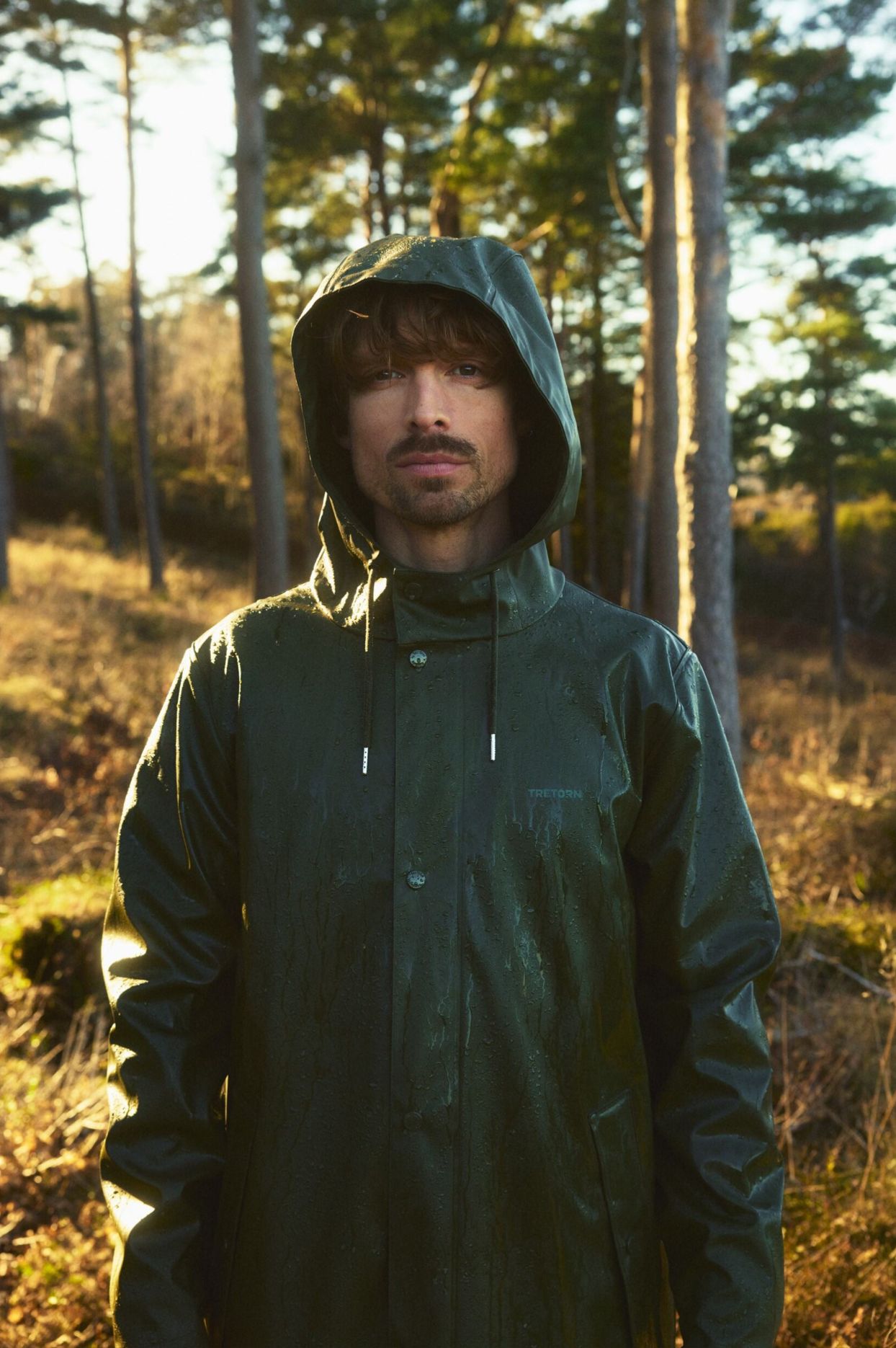A person in a green raincoat stands in a sunlit forest, with trees and dry grass in the background.