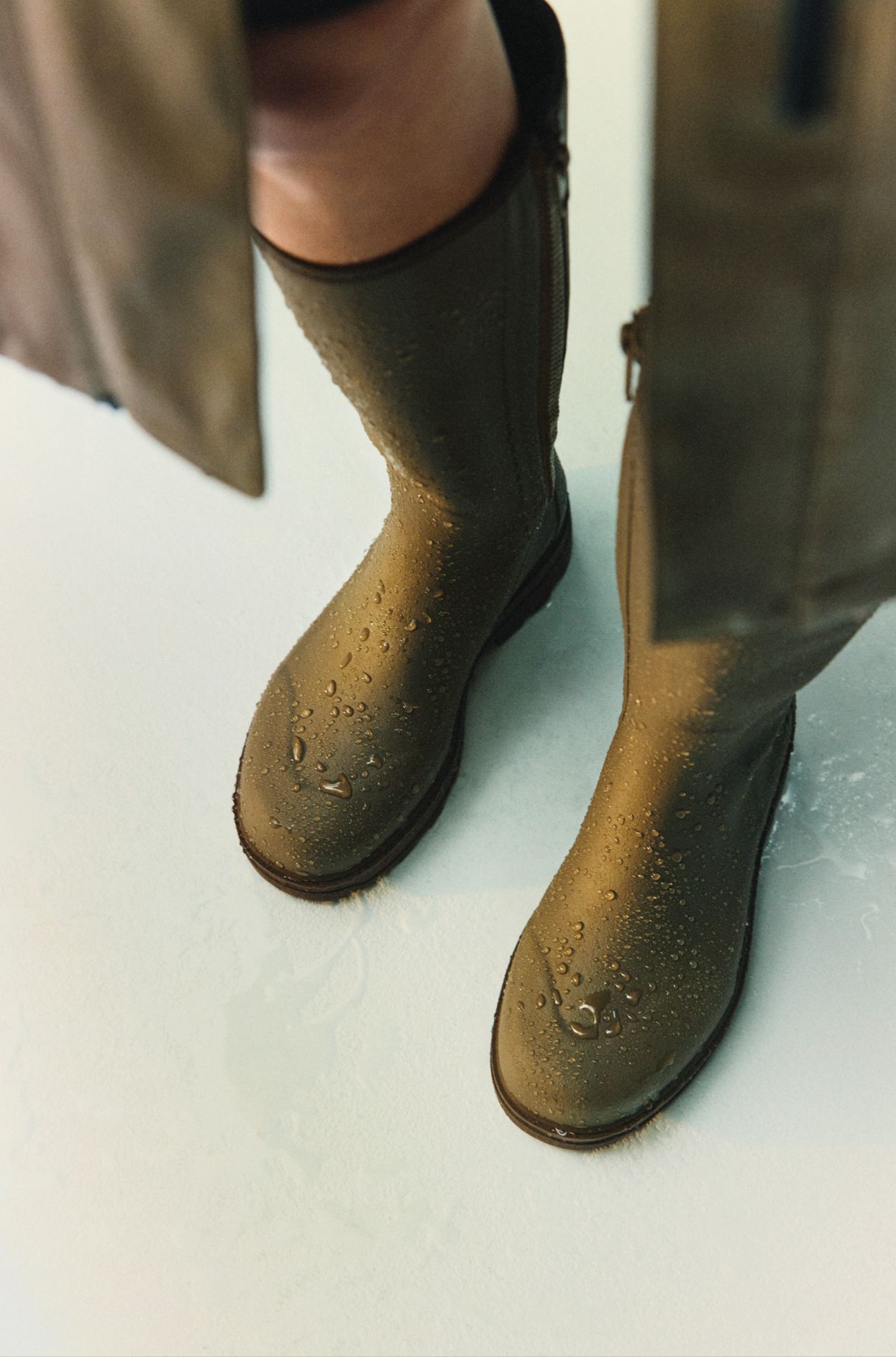 Person wearing brown rain boots with water droplets, standing on a wet surface. A coat is partially visible.