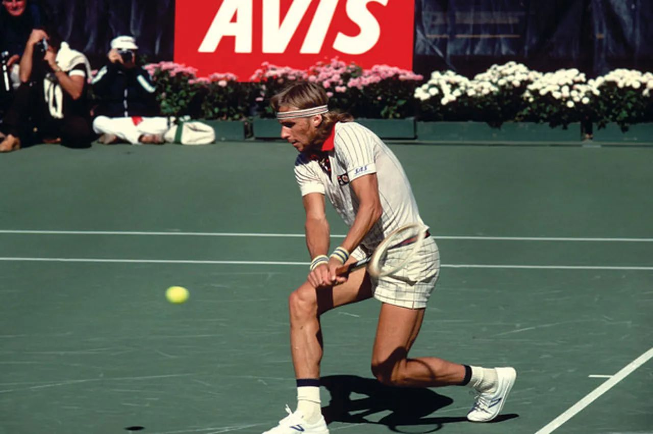 Tennis player in action, wearing a headband and striped outfit, hitting a backhand shot on a green court with an AVIS sign in the background.