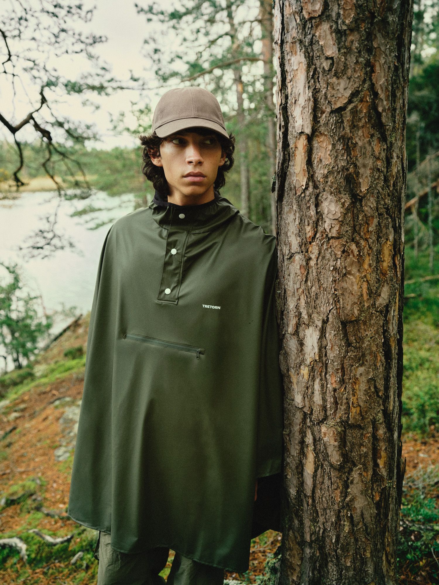 Person in green poncho and cap stands beside a tree in a forest, with a lake and greenery in the background.