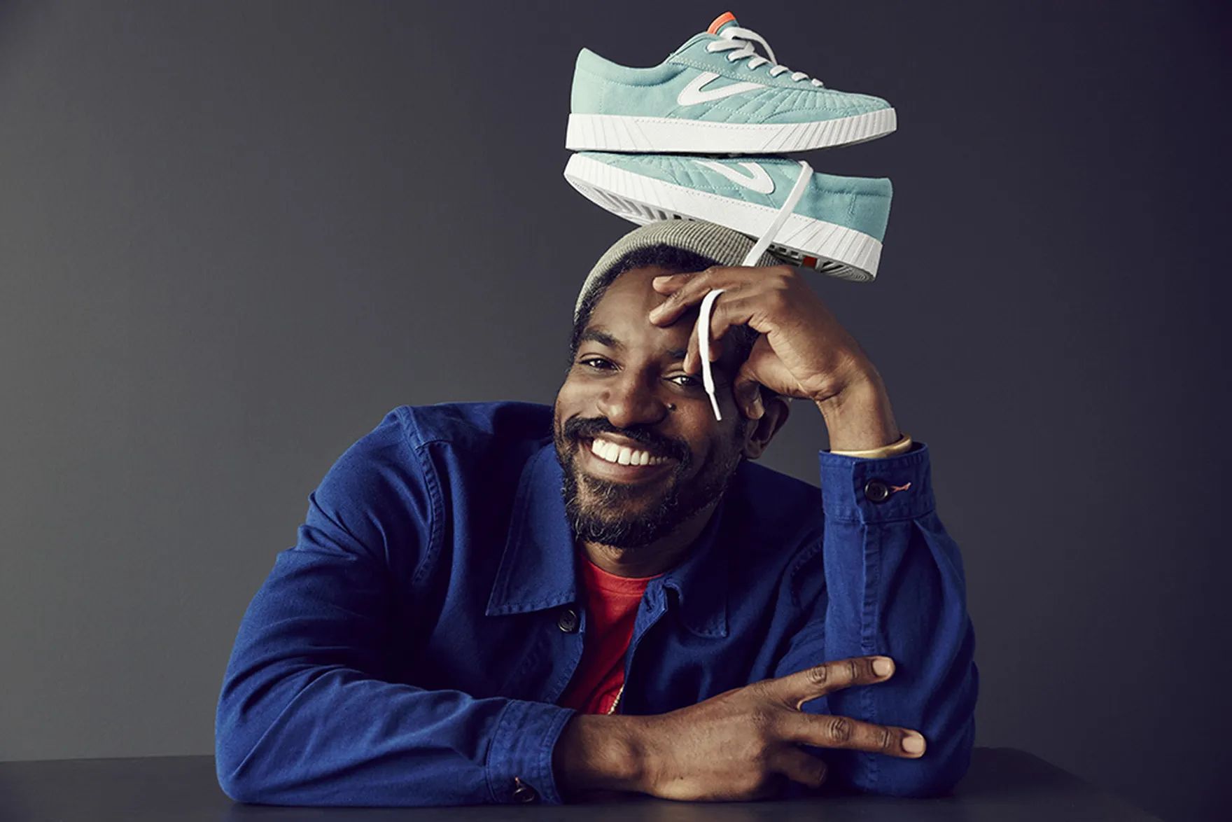 Man smiling, wearing a blue jacket and beanie, with turquoise sneakers balanced on his head against a dark background.