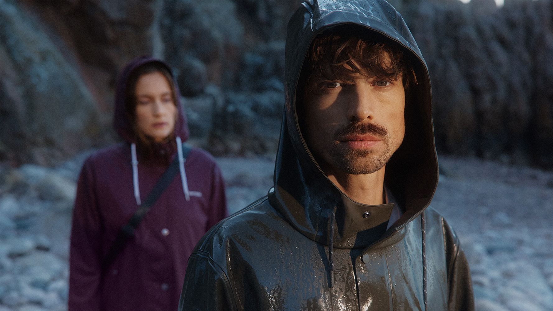 Two people in raincoats stand outdoors; the person in front wears a shiny black coat, and the one behind wears a purple coat.