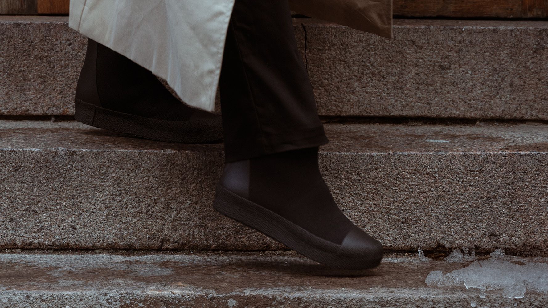 Person in dark shoes and pants, wearing a beige coat, walking down stone steps with a small patch of ice nearby.