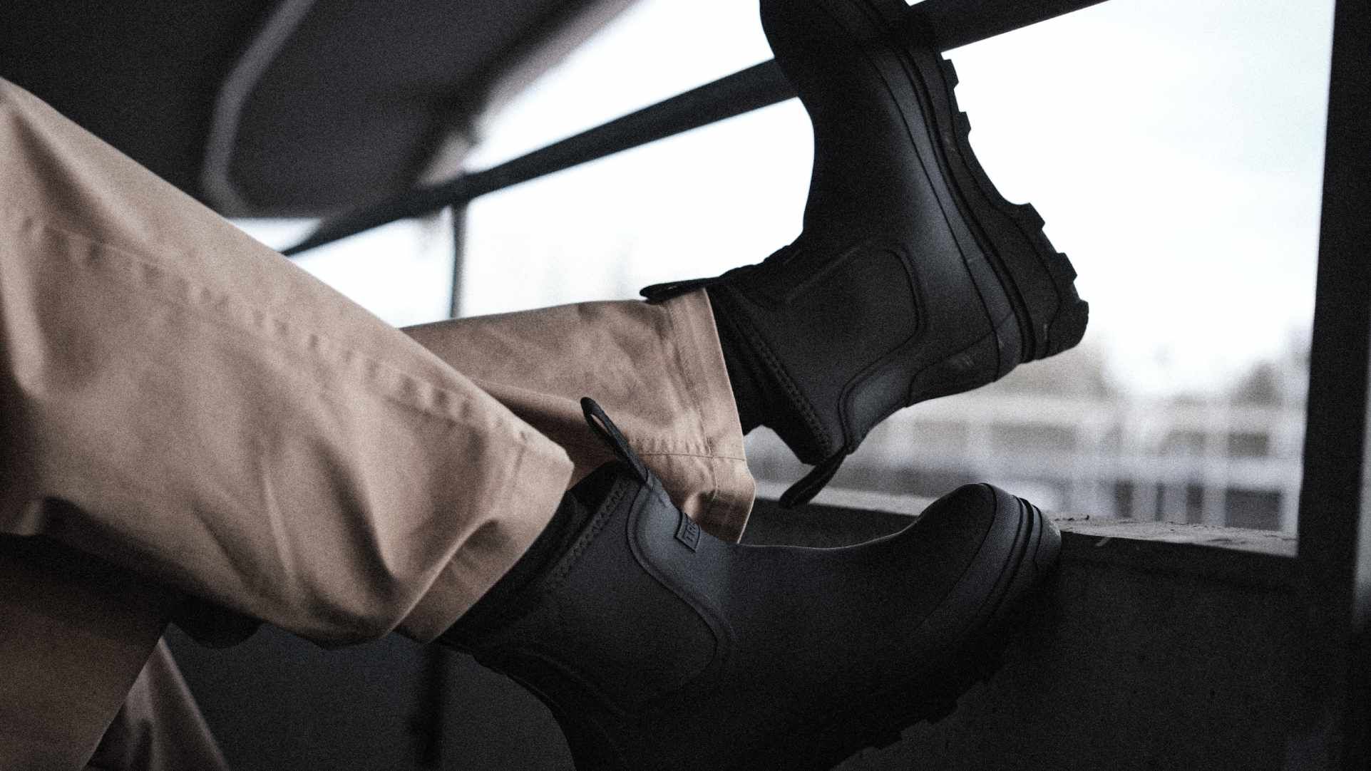 Person wearing tan pants and black boots, resting feet on a ledge, with an industrial background and overcast sky.