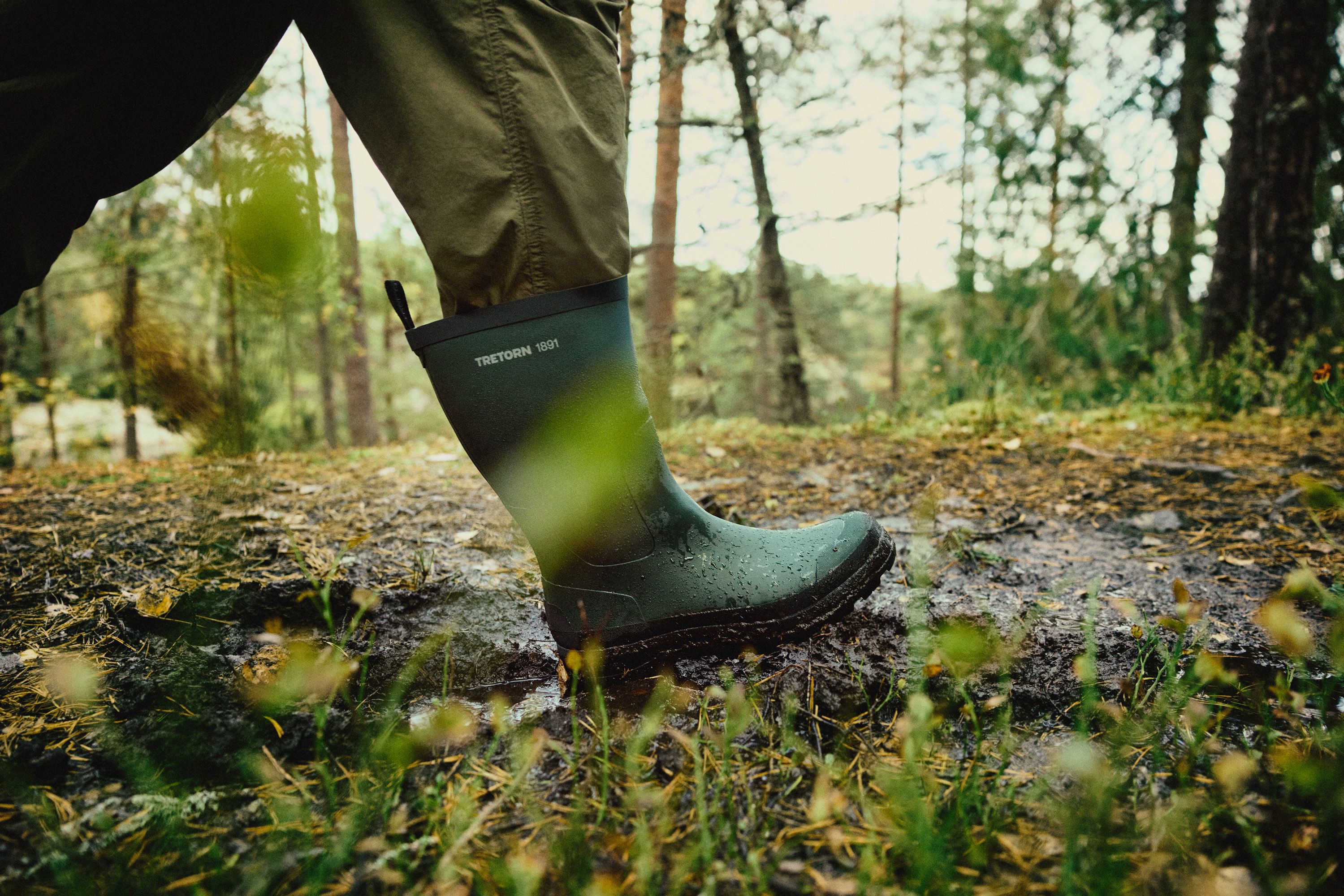 Close-up of a person wearing green rubber boots walking through a muddy forest trail, surrounded by trees and greenery.