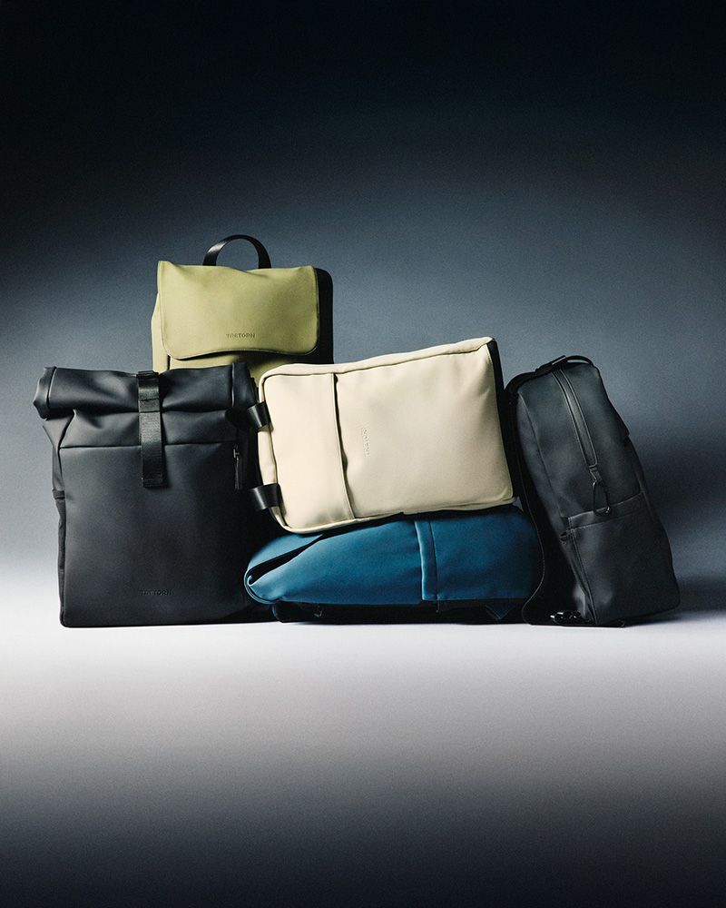 A collection of stylish bags, including backpacks and duffels, in black, beige, green, and blue, arranged against a dark gradient background.