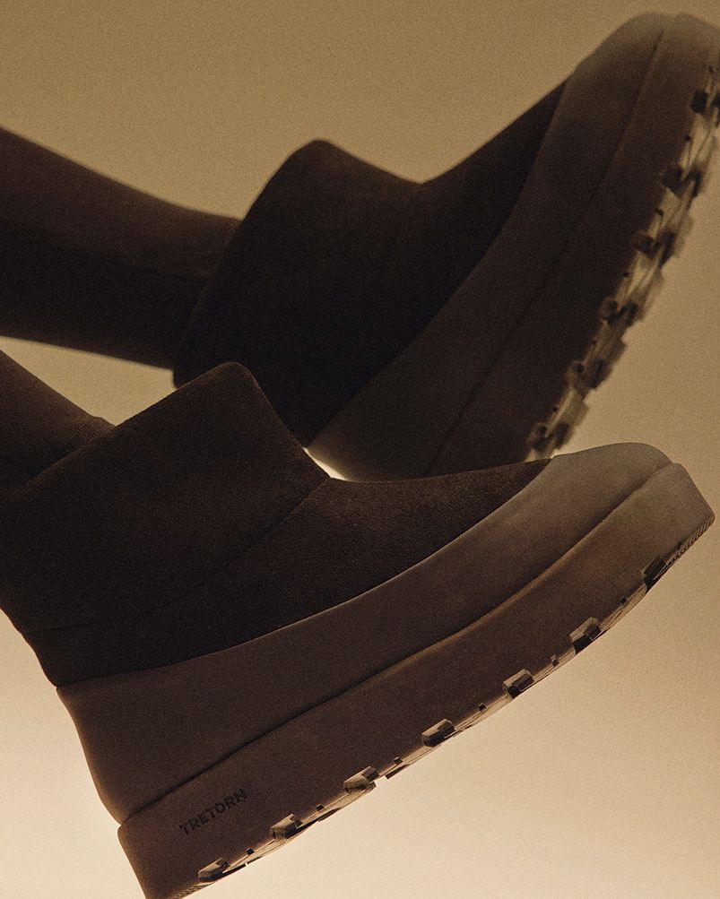 Close-up of dark brown boots with thick soles and textured treads, angled against a warm, softly lit background.